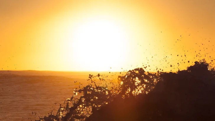 Yellow sky with the white colored sun during an ocean sunset with a wave crashing up